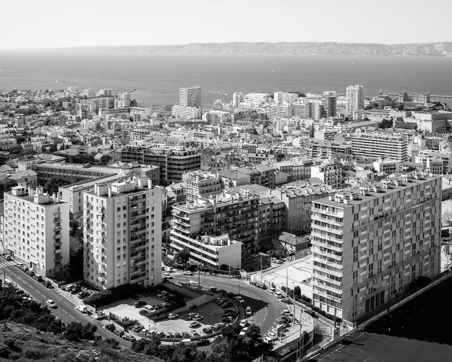 Aerial black and white cityscape with dense urban buildings overlooking a calm sea horizon.
