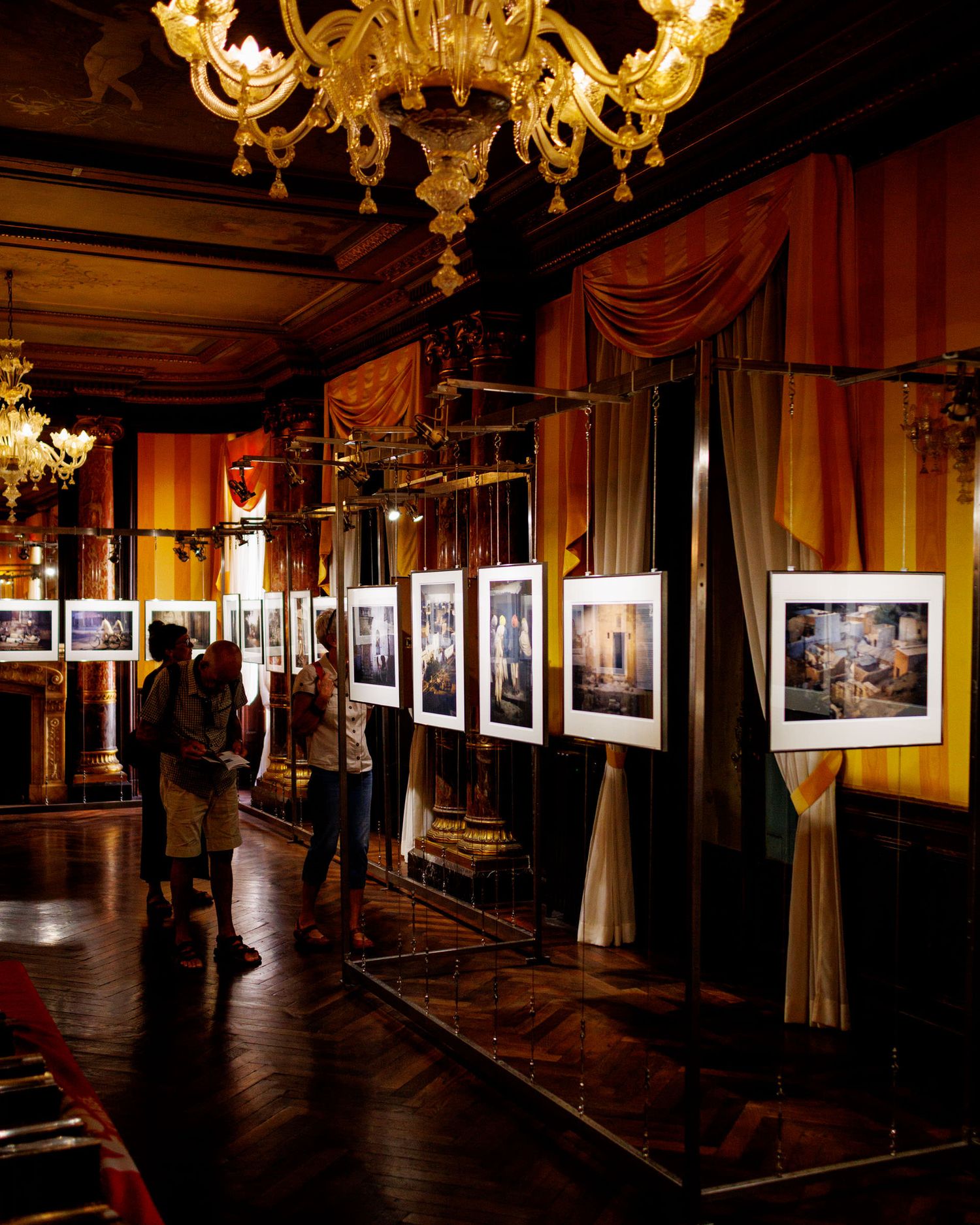 Visitors browsing framed photographs in an opulent gallery room with golden chandeliers and draped walls.