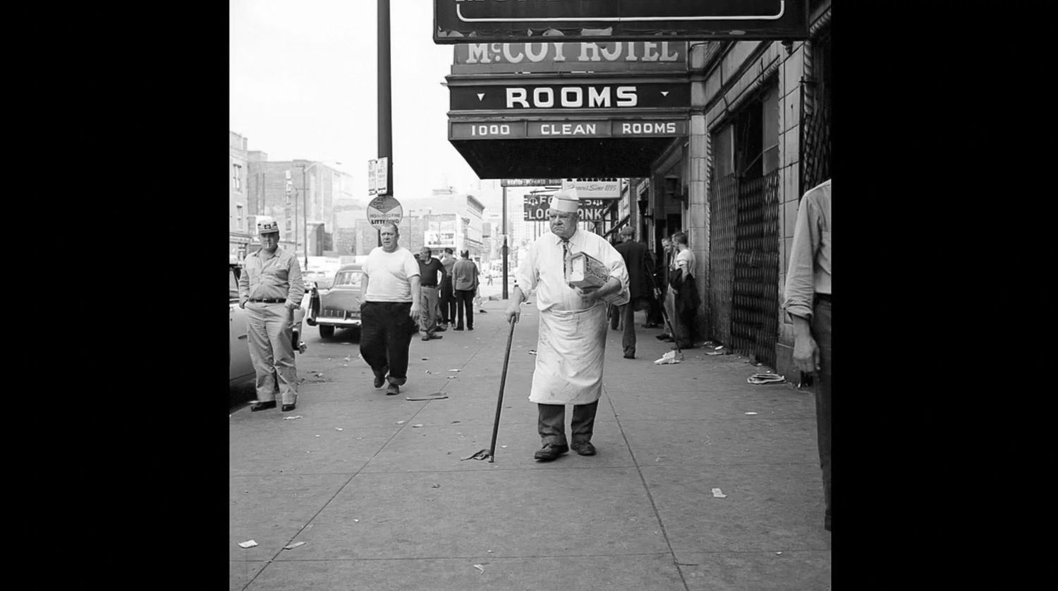 Black and white street photo of a man in a white apron holding a cane on a city sidewalk beneath a vintage hotel rooms sign.