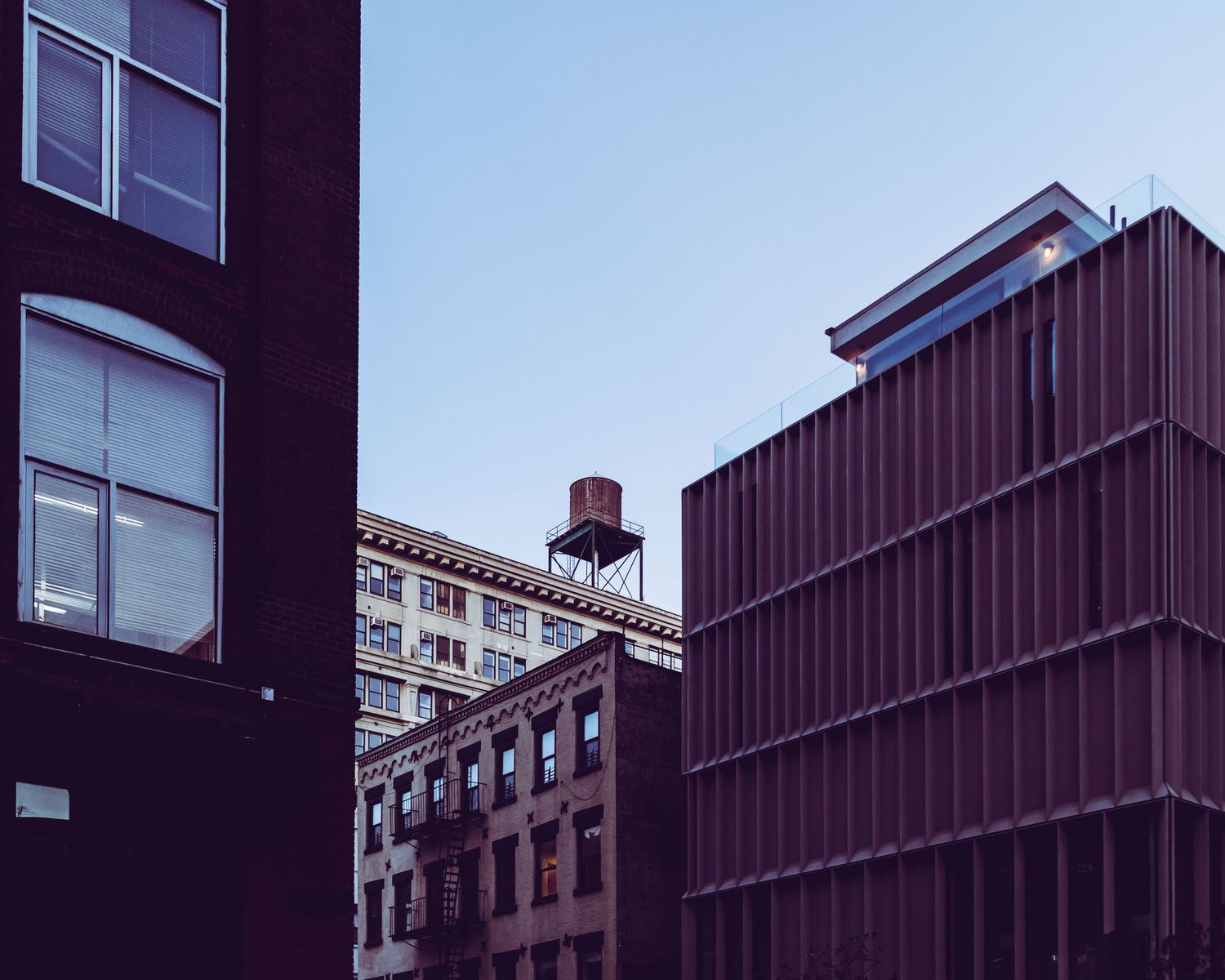 Urban cityscape of brick and metal buildings against a clear blue sky with a rooftop water tower visible.