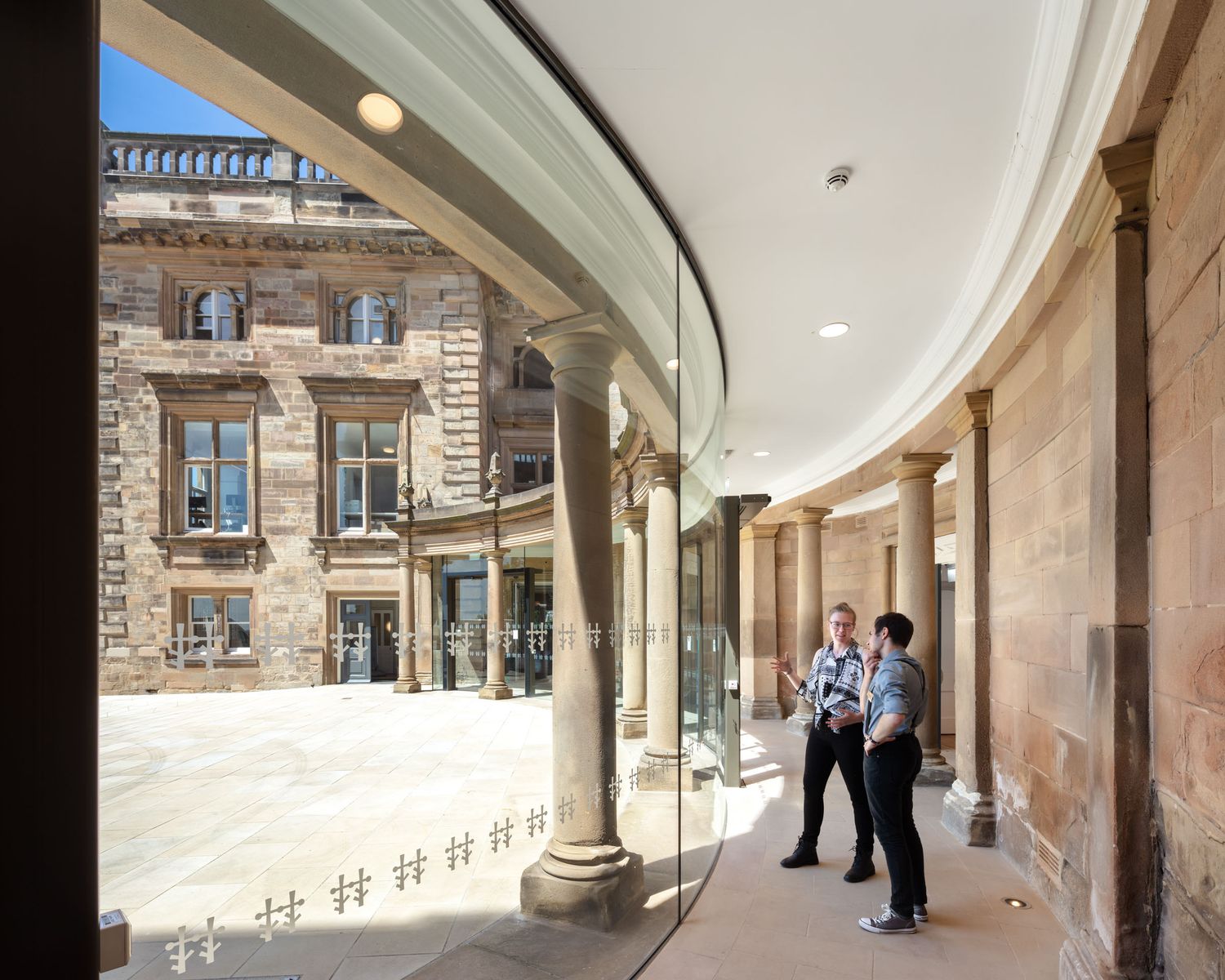 Curved glass colonnade connecting historic sandstone buildings in a renovated courtyard.