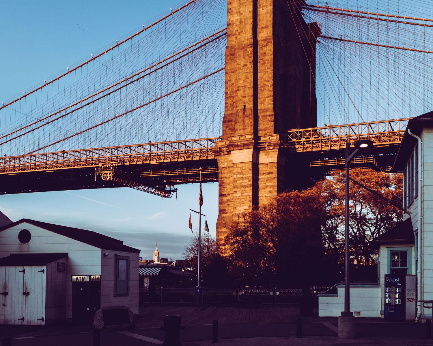 Golden-lit Brooklyn Bridge tower looms over waterfront buildings at dusk with a clear blue sky backdrop.