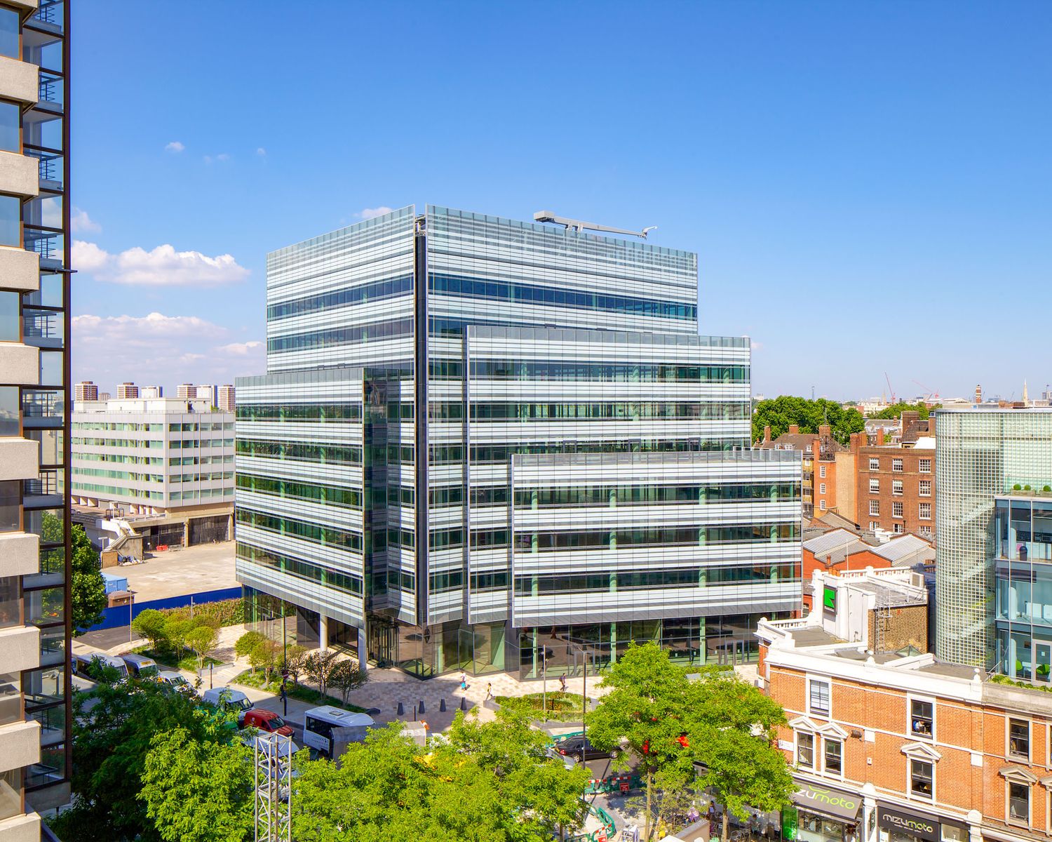 Modern glass office building with multiple floors surrounded by urban streets and green trees under a clear blue sky.