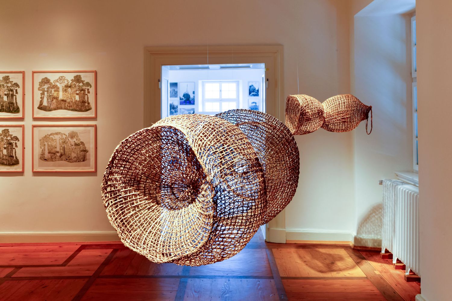Large woven basket sculpture suspended in a gallery with framed artwork on walls and warm red carpet flooring.