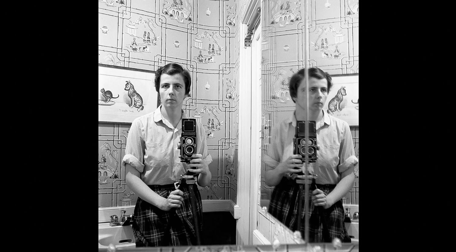 Black and white mirror selfie of a person holding a twin-lens reflex camera, standing in a bathroom with cat-print wall tiles.