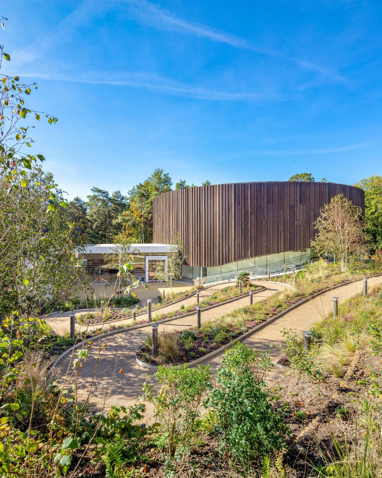 Modern timber-clad building surrounded by landscaped gardens and native planting under a bright blue sky.