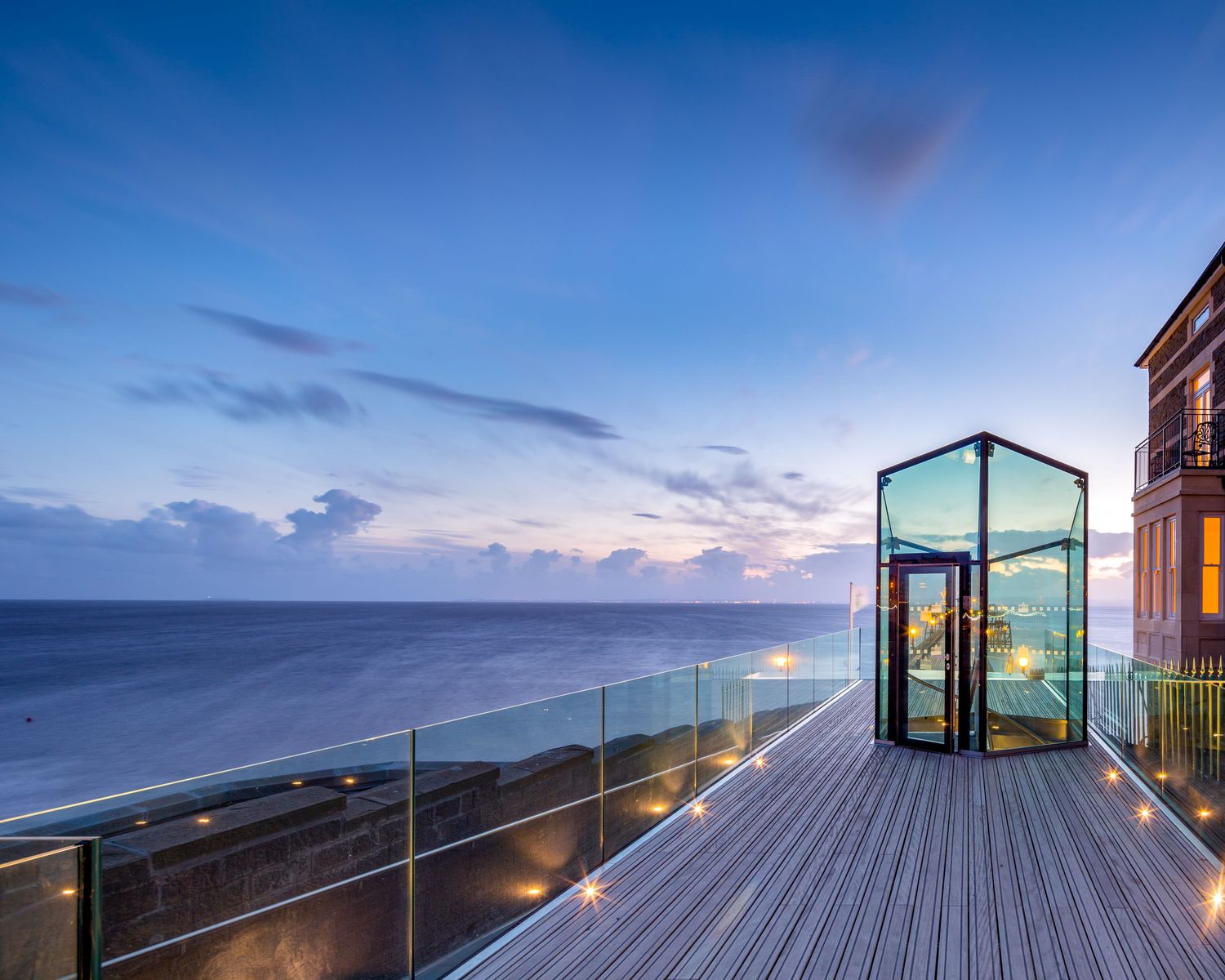 Glass elevator on a wooden deck overlooking a serene ocean at dusk with glowing lights and a blue sky.