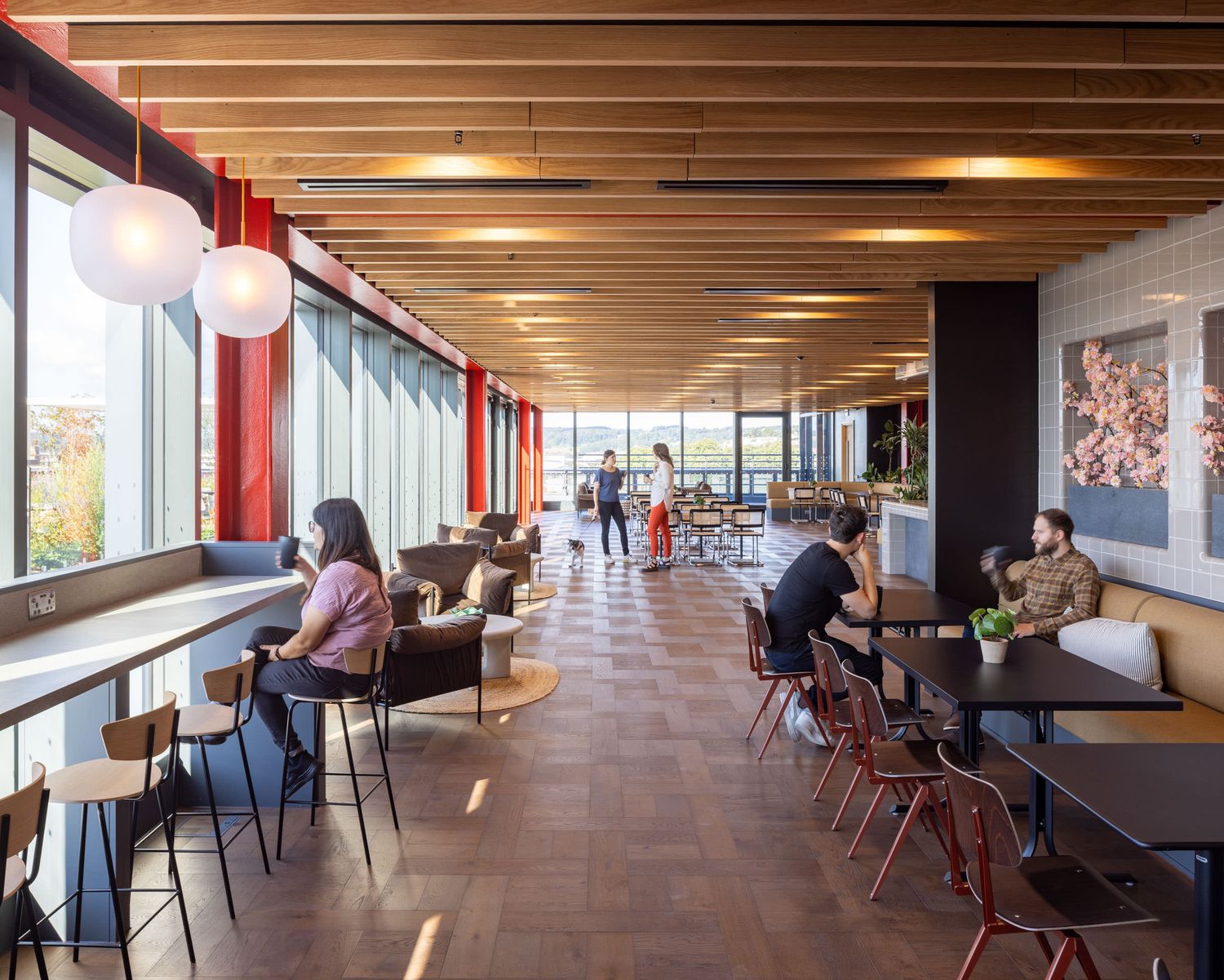 Modern restaurant lounge with wood ceiling, pendant lights, large windows, and patrons seated at tables.