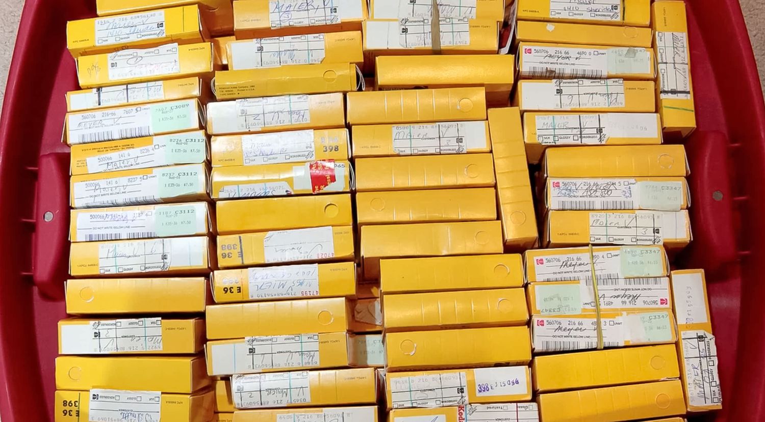 Red plastic tray filled with stacked yellow and white film boxes, showing vintage photographic film packaging in bulk.