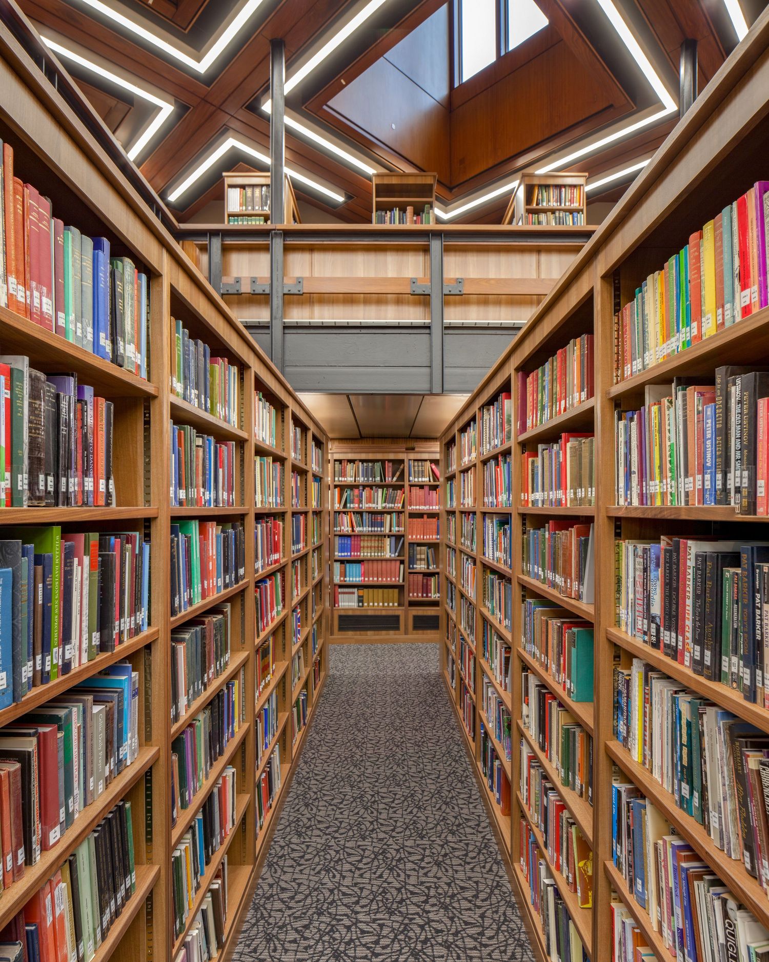A symmetrical library aisle flanked by tall wooden shelves filled with colorful books under modern skylights.