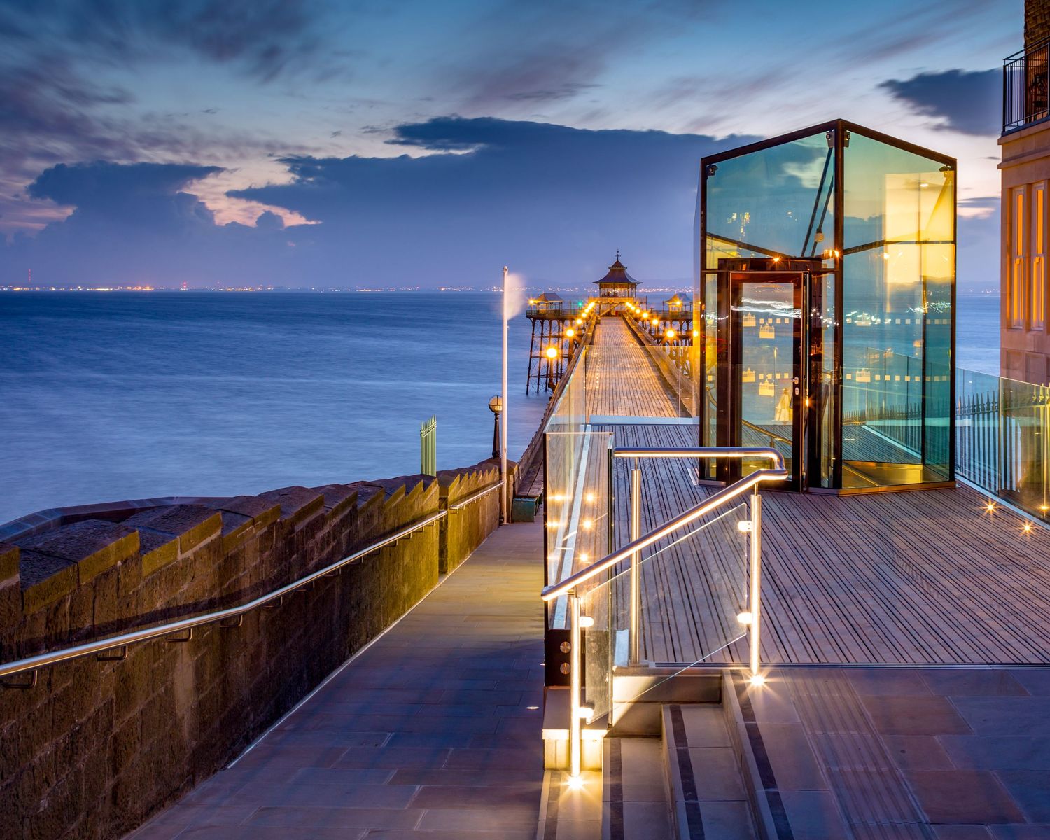 Glass elevator and illuminated pier extending over the ocean at dusk with glowing lights reflecting on the boardwalk.