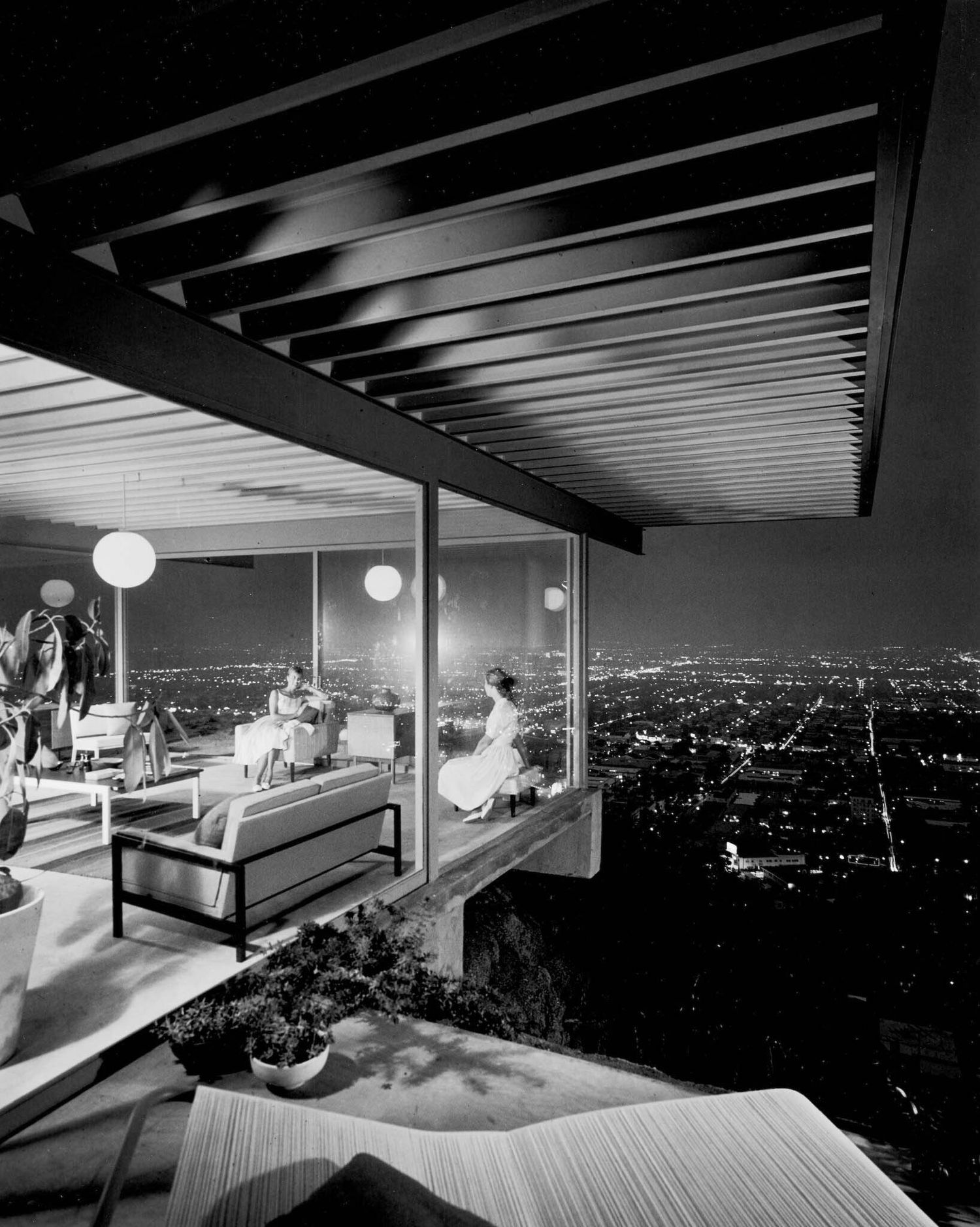 Modernist hillside home at night with panoramic city lights view, featuring open terrace and mid-century furniture.