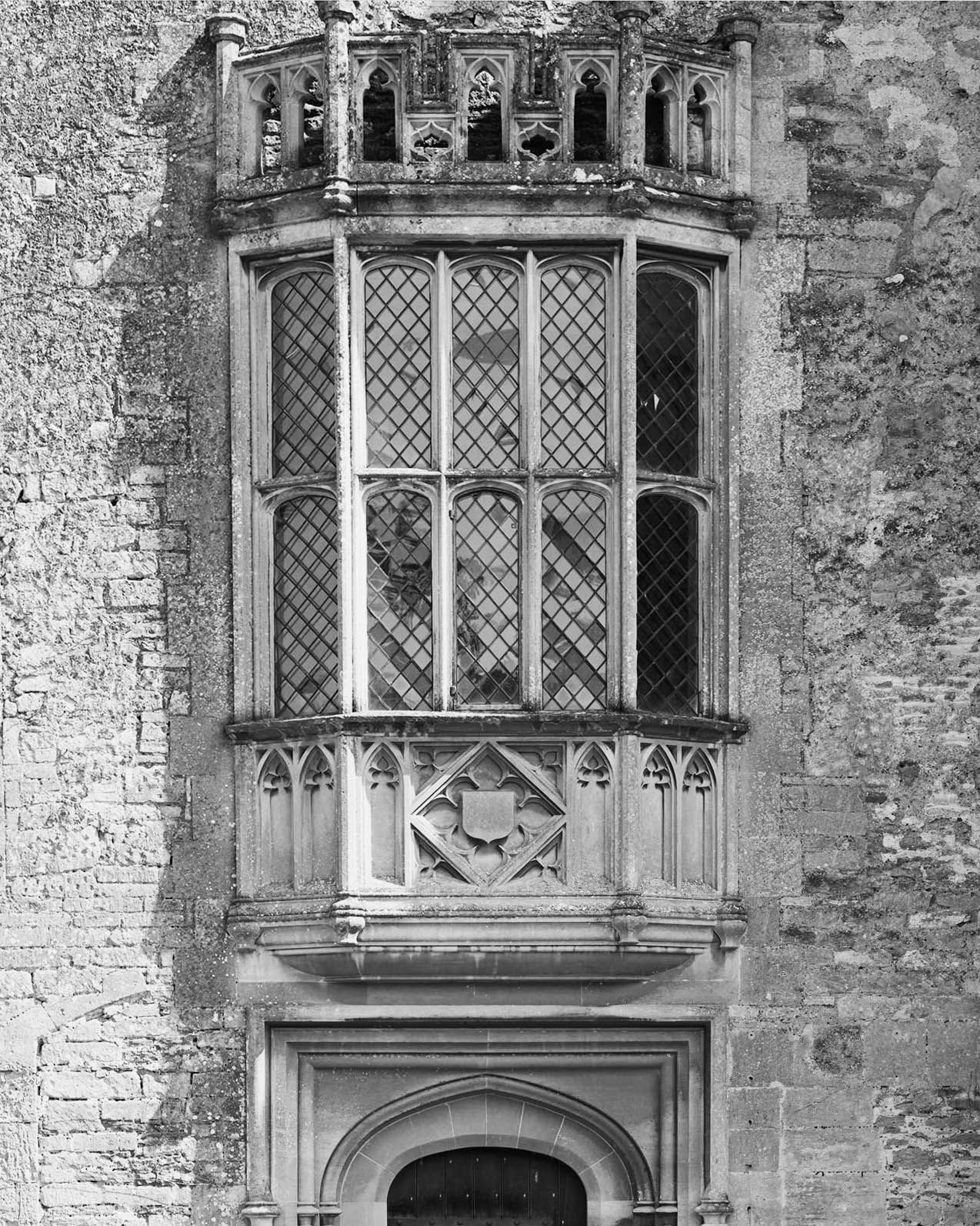 Black and white photo of a Gothic stone bay window with leaded glass above an arched doorway on a historic building.