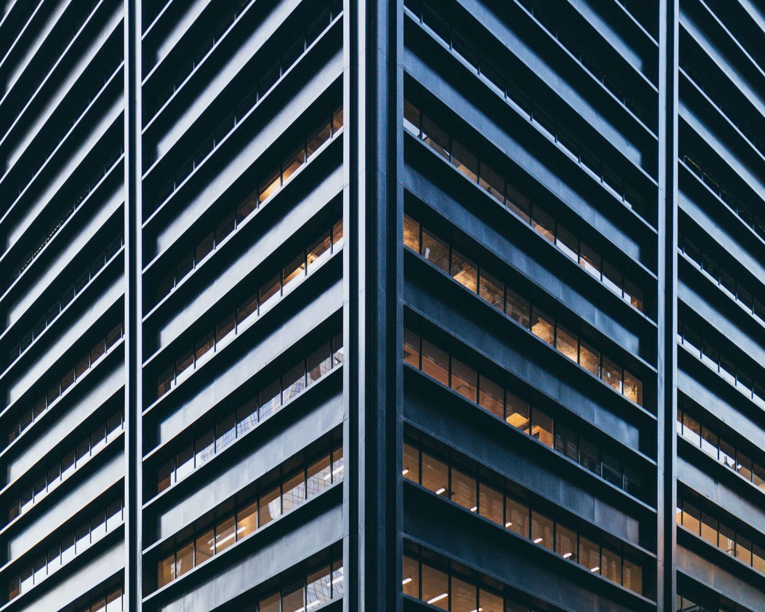 Modern glass skyscraper corner with illuminated office windows reflecting blue steel facade at dusk.