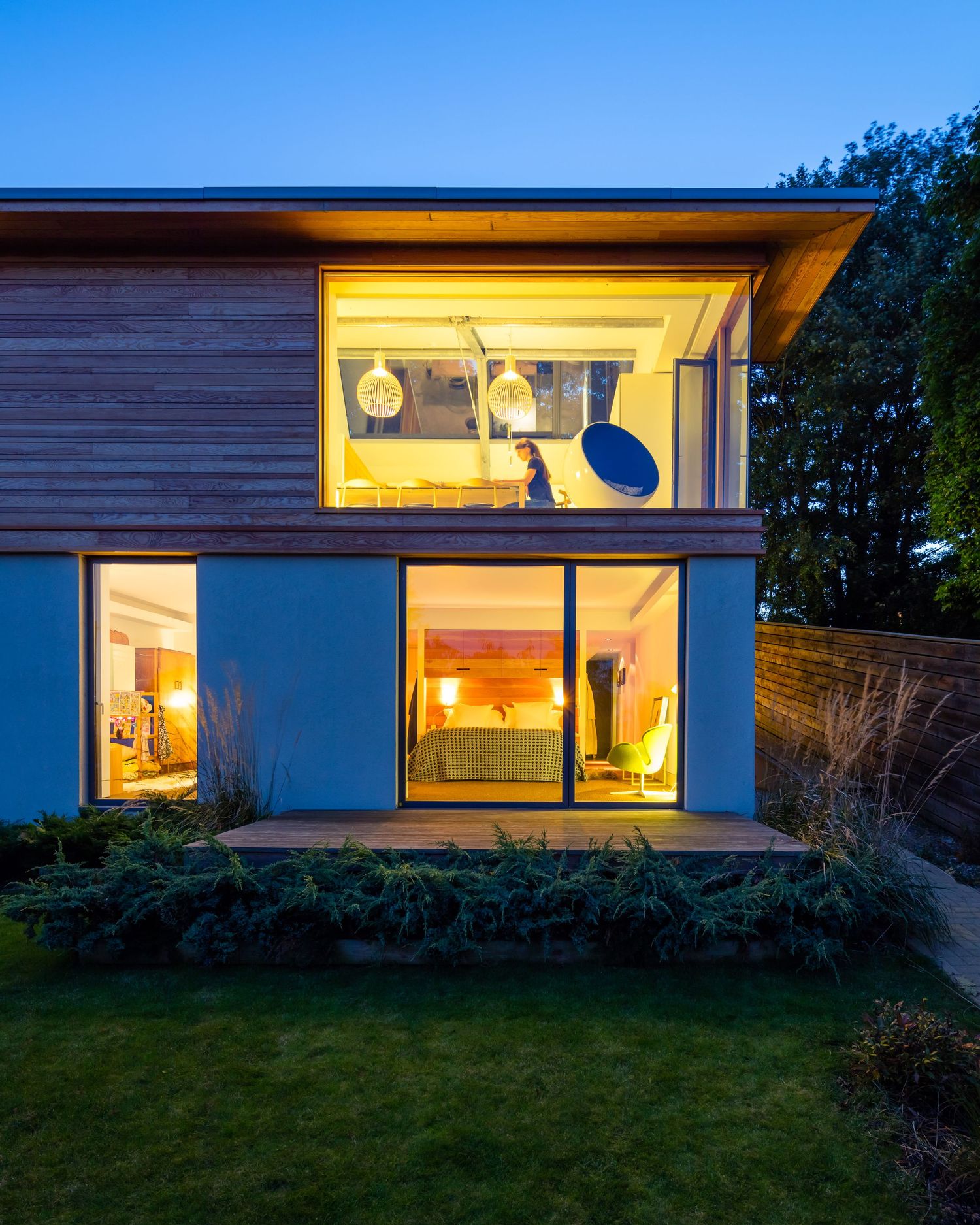 Modern two-story home exterior at dusk with warm glowing interior lights, large windows, wood cladding, and lush green lawn.