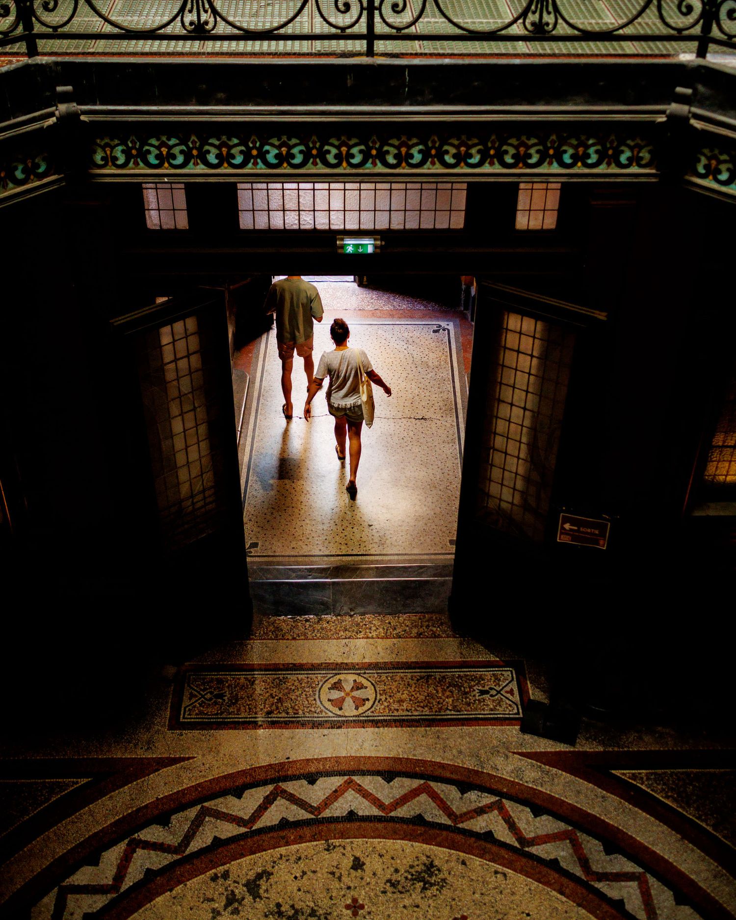 Two people walking through an ornate tiled corridor with decorative mosaic floors in a historic building.