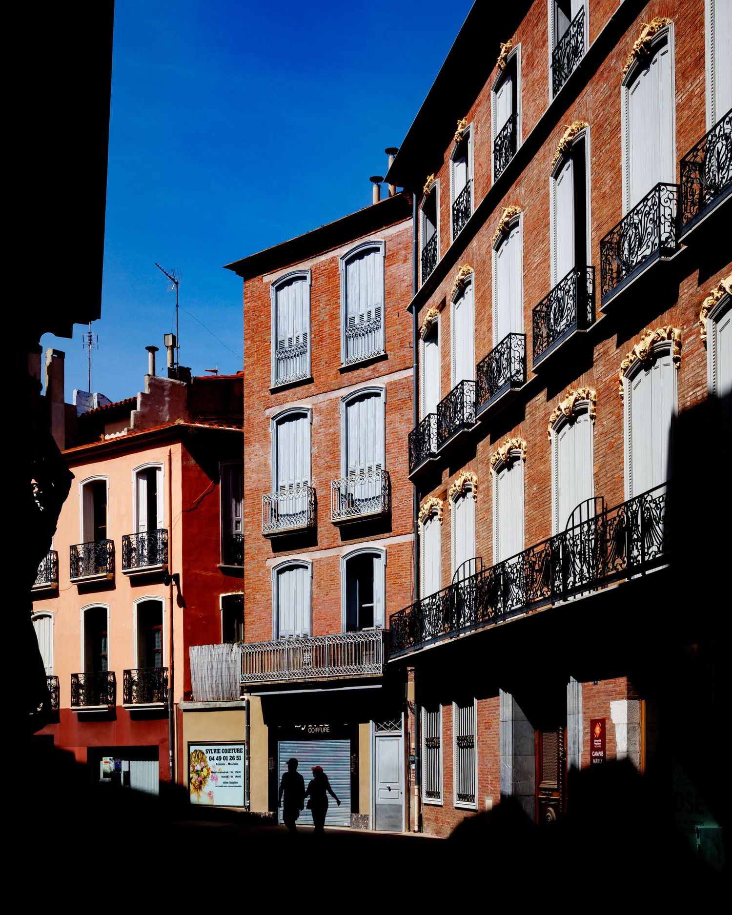 Sunlit brick apartment buildings with ornate iron balconies casting dramatic shadows on a European street.