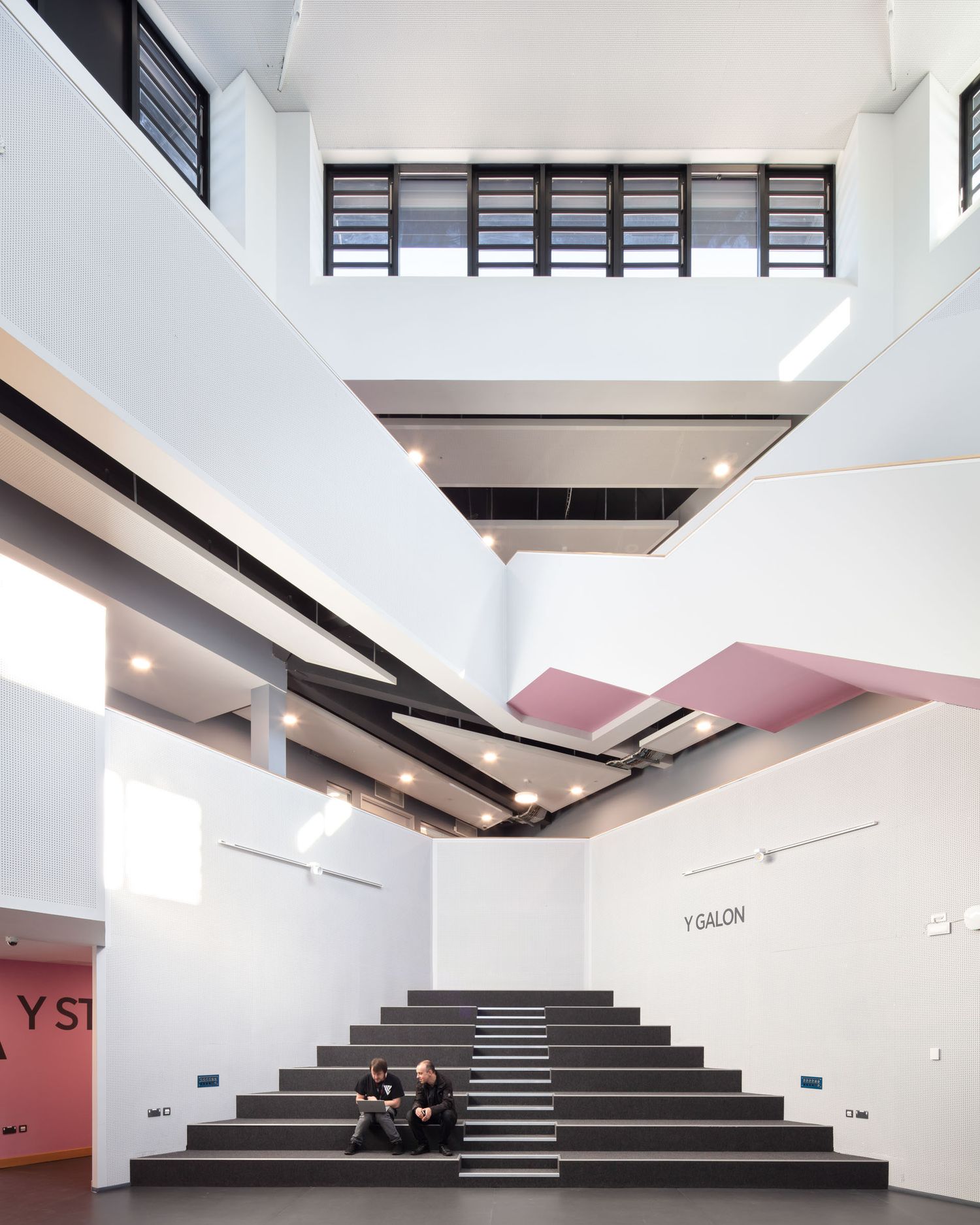 Modern atrium with gray stairs, white walls, pink accents, and two people sitting, labeled Y Galon.