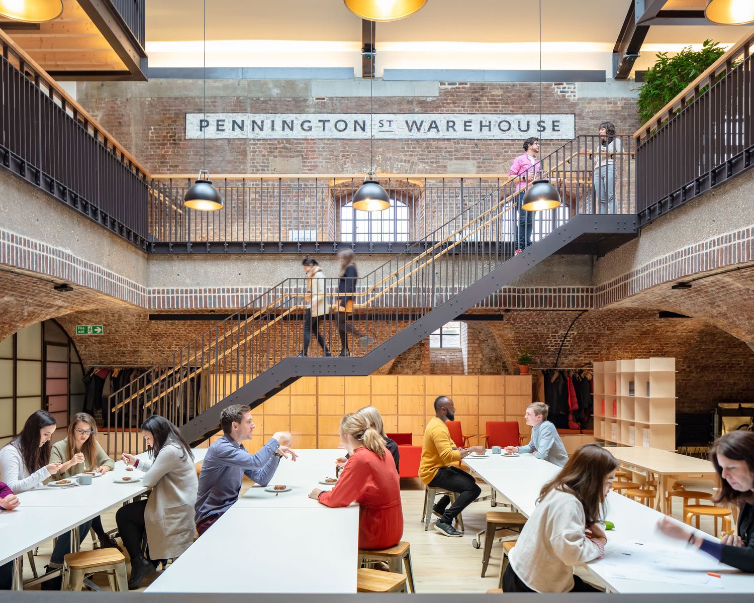 Modern collaborative workspace inside Pennington Street Warehouse with brick walls, staircases, and people working at communal tables.