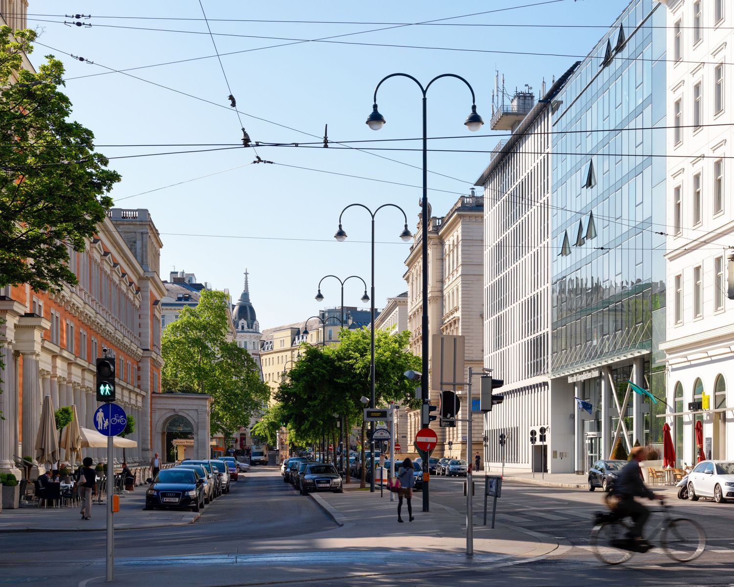A sunny urban street in Vienna with cyclists, parked cars, green trees, and modern glass building alongside classic architecture.