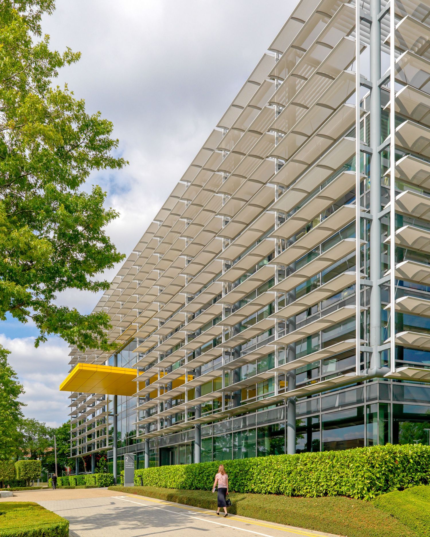 Modern glass office building with solar shading panels, green hedges, and a pedestrian walkway on a sunny day.