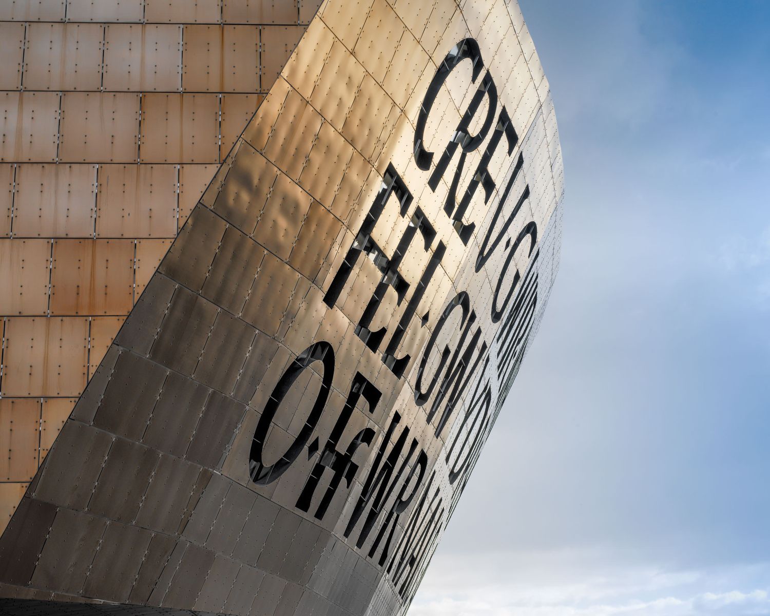 Close-up of the Wales Millennium Centre facade with Welsh text inscription against a cloudy sky.