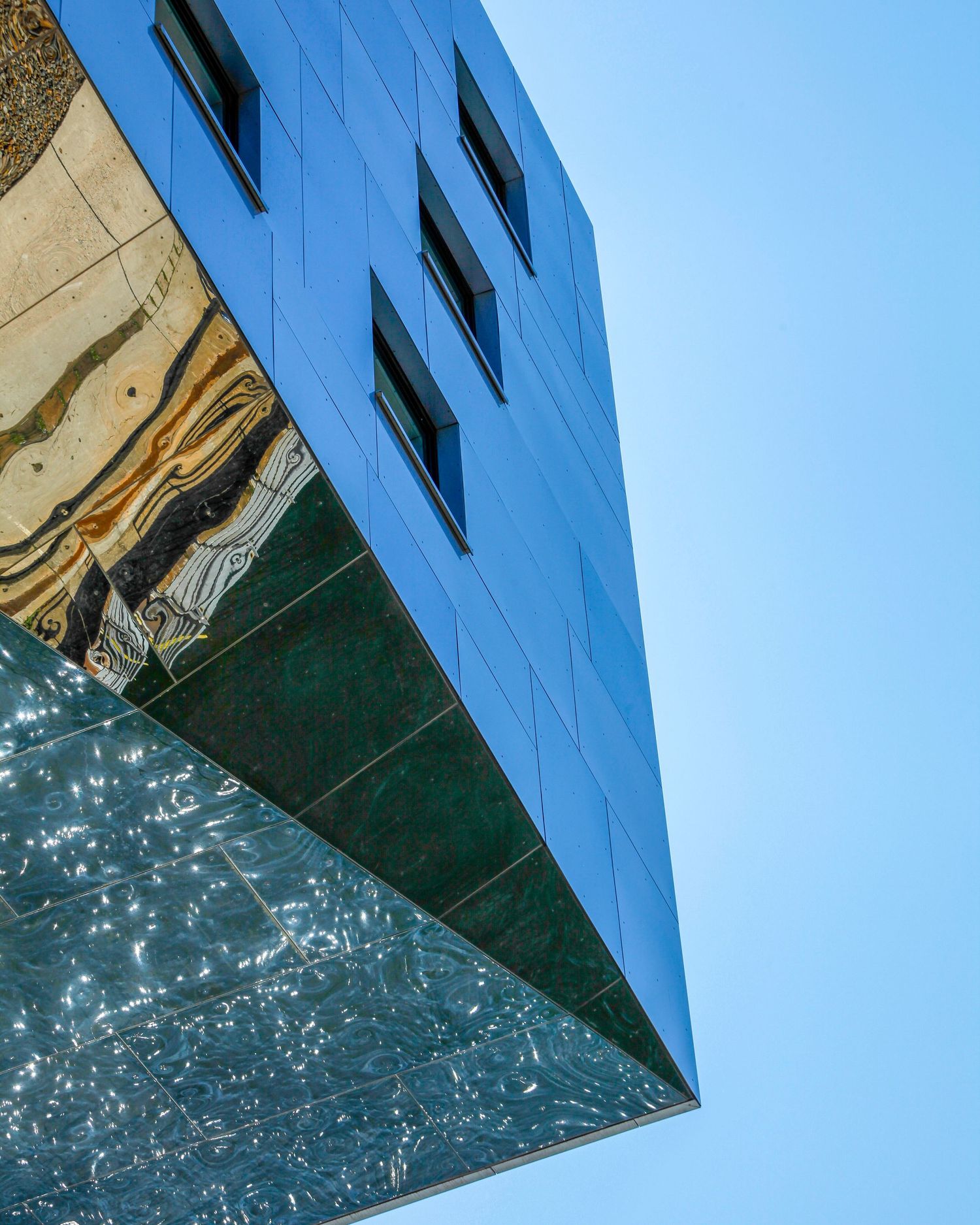 Low-angle view of a modern blue glass building with reflective marble panels against a clear blue sky.