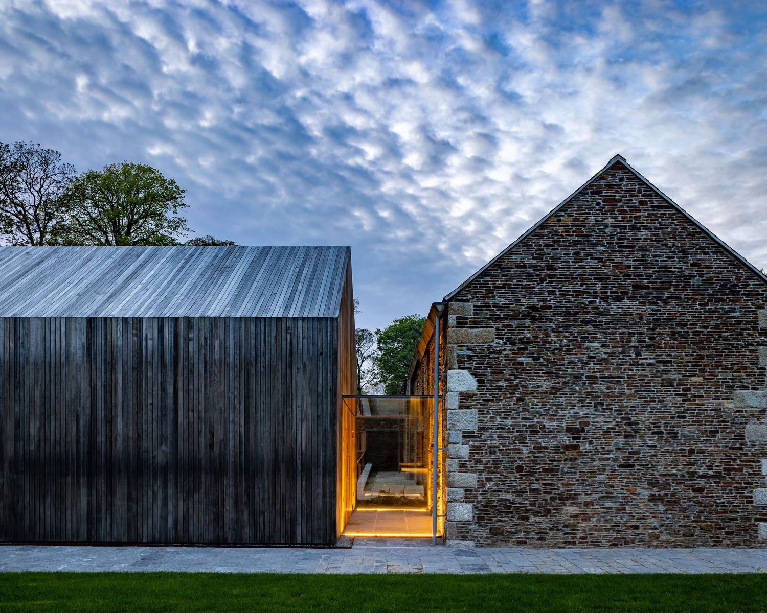 Modern barn conversion with dark timber cladding beside a stone building at dusk, dramatic cloudy sky.