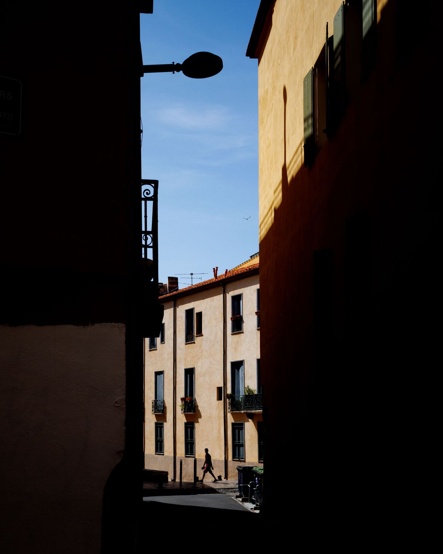 Narrow alleyway between shadowed buildings opening to sunlit street with pedestrian and classic architecture.