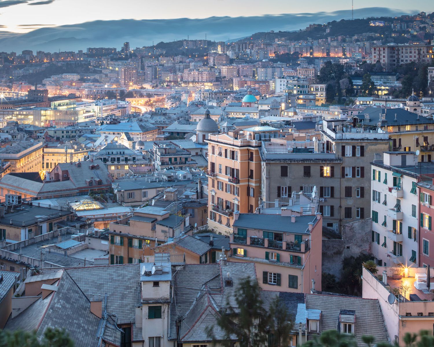 Aerial twilight view of densely packed historic colorful buildings across a hillside cityscape in Genoa, Italy.