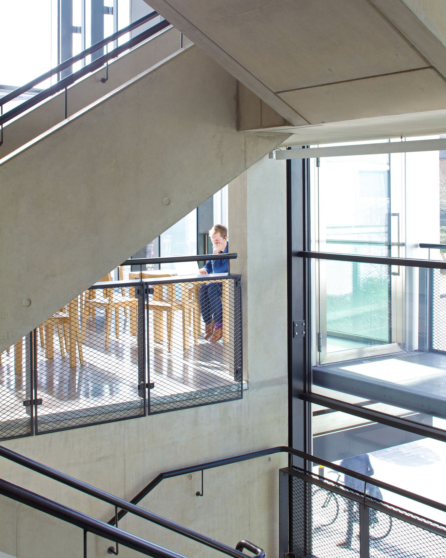 Modern concrete staircase with metal mesh railings and a person standing on an upper floor mezzanine level.