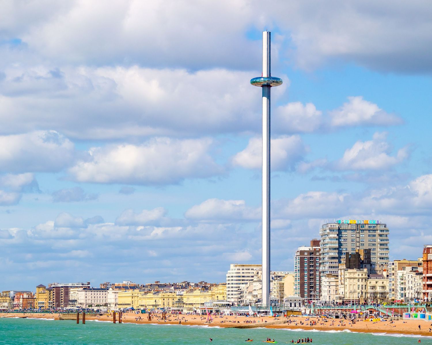 Brighton seafront with the British Airways i360 observation tower rising above the beach and colorful buildings under a cloudy sky.