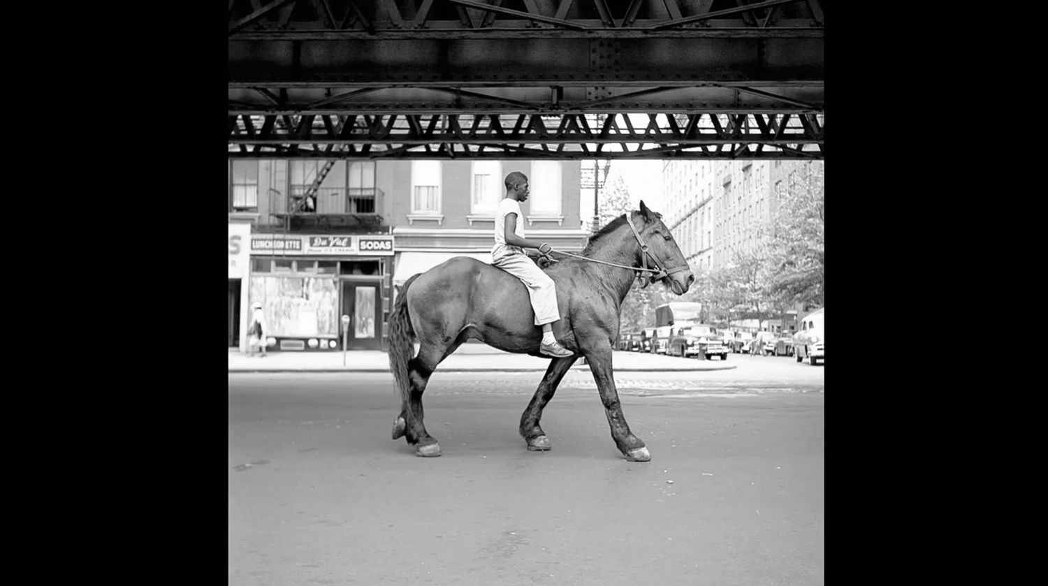 Black and white photo of a person riding a horse down an empty city street beneath an elevated steel train bridge.