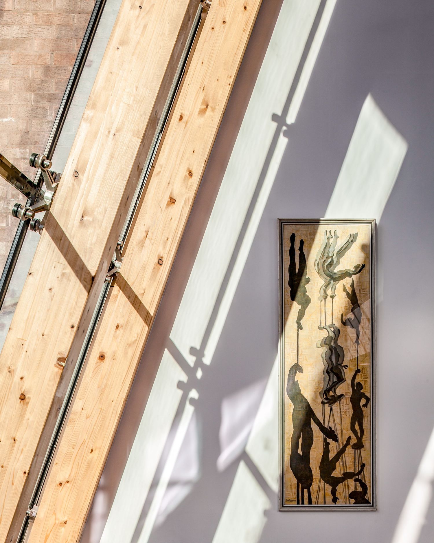 Framed artwork with human figures displayed on a sunlit wall beside wooden beams and metal rods in a modern interior.