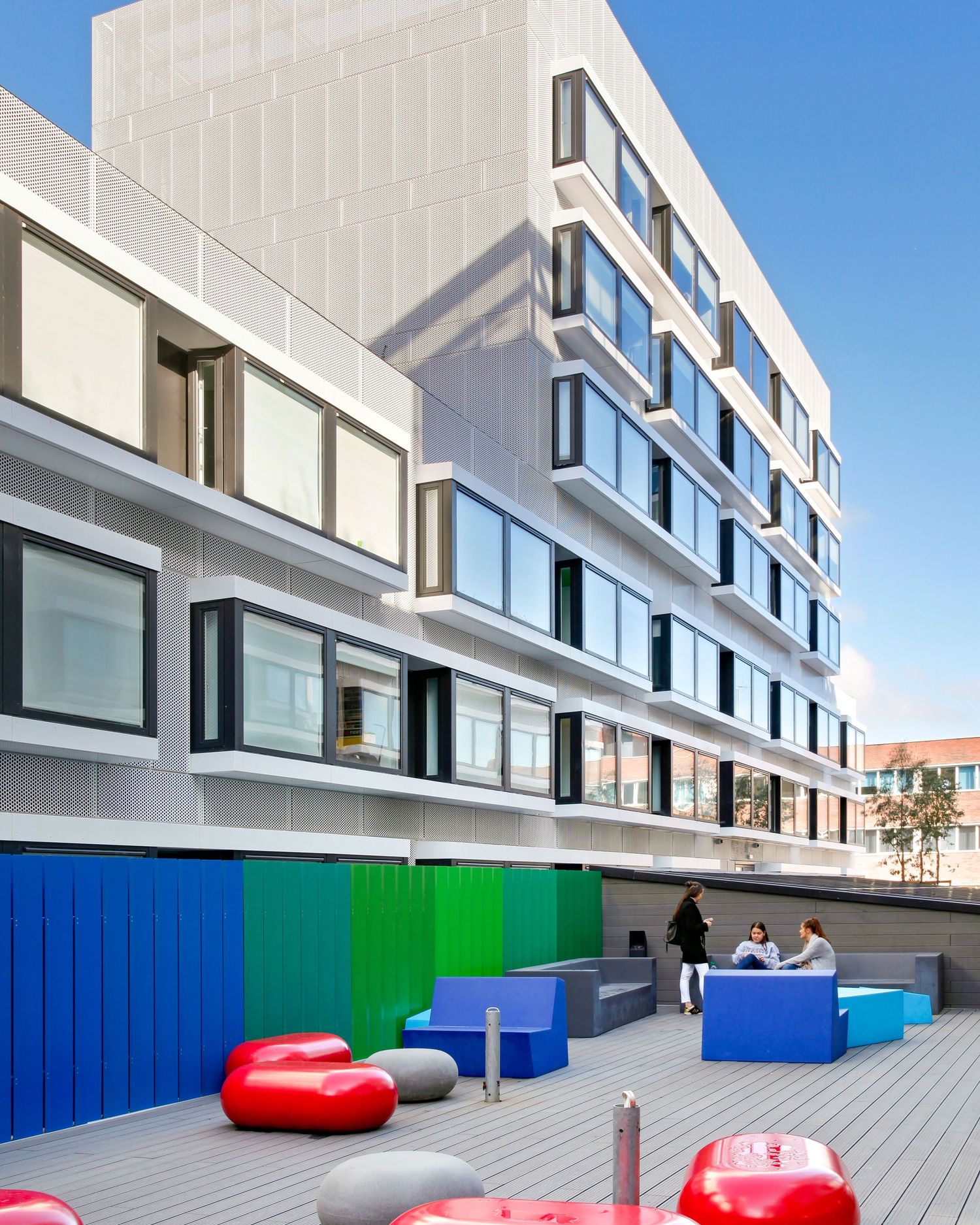 Modern building rooftop terrace with colorful blue, green, red seating and gray decking, people socializing outdoors.