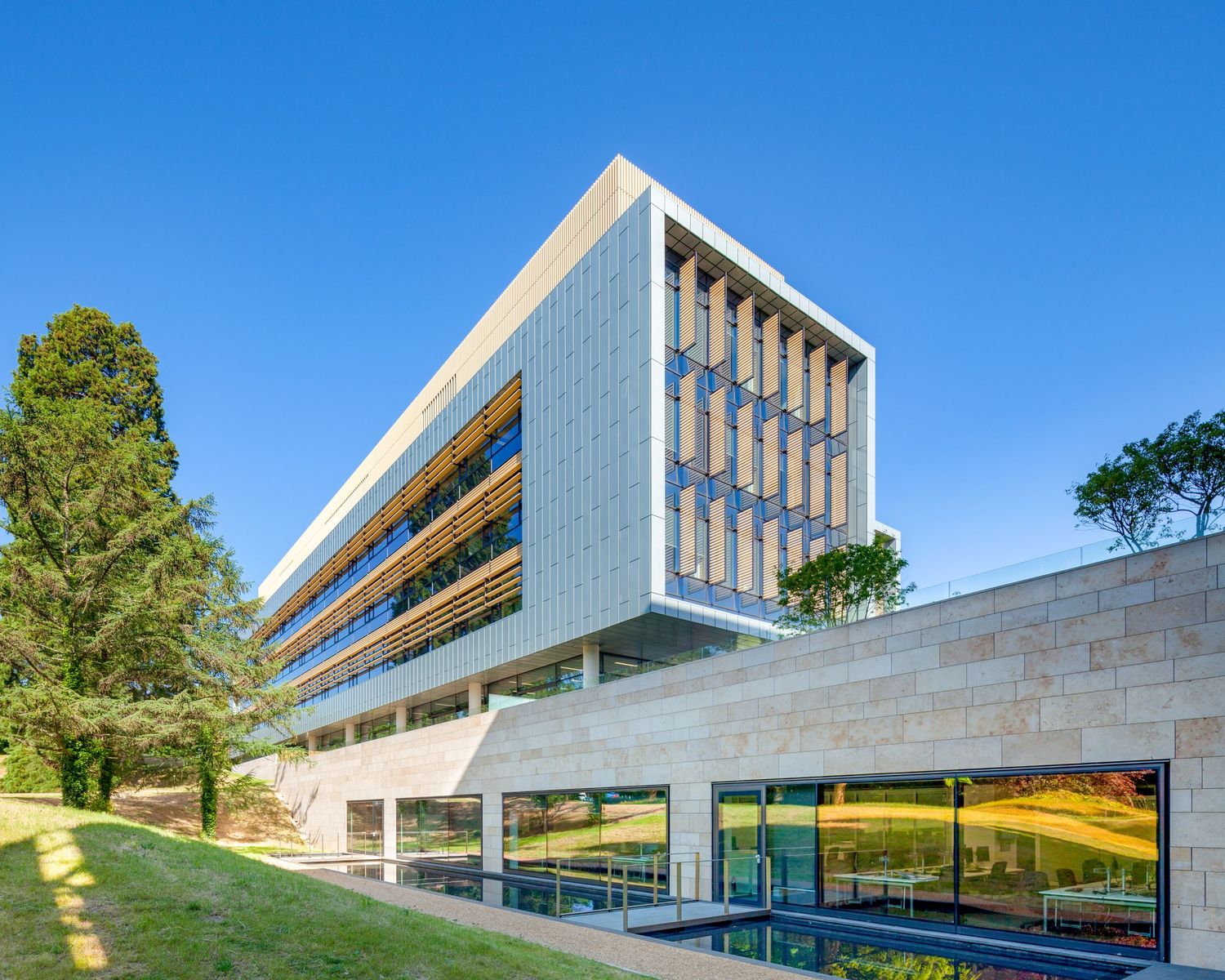 Modern multi-story office building with wood panel facade, glass windows, and landscaped green surroundings under blue sky.