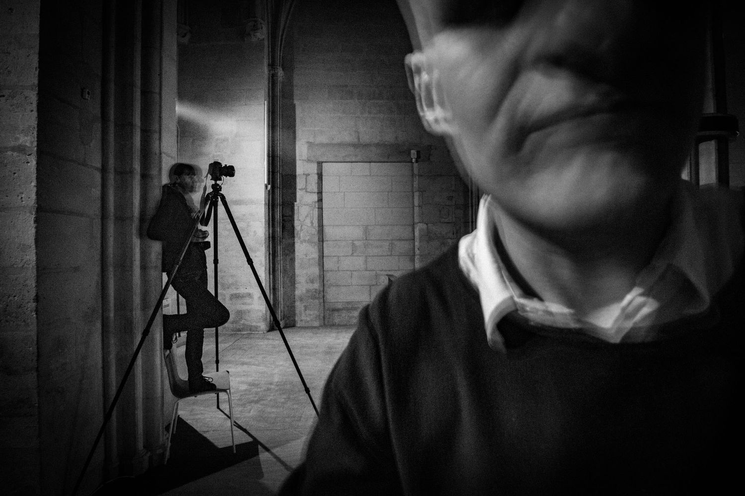 Blurred man in foreground with photographer on tripod in dark industrial space.
