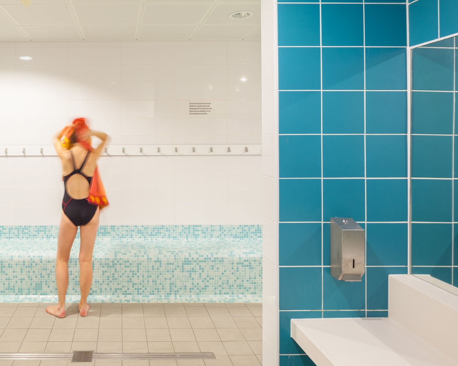 Woman in black swimsuit adjusting hair near blue tiled shower wall in modern pool changing room.