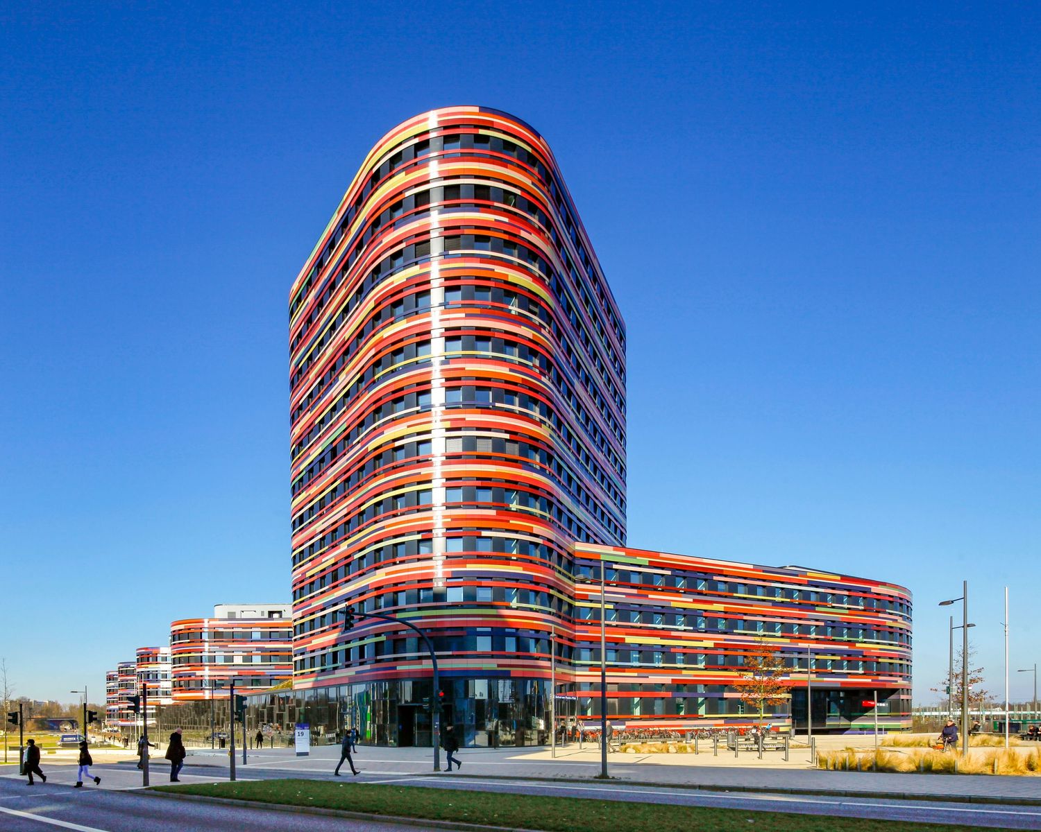 Modern curved red and white high-rise building with horizontal balconies under a clear blue sky.