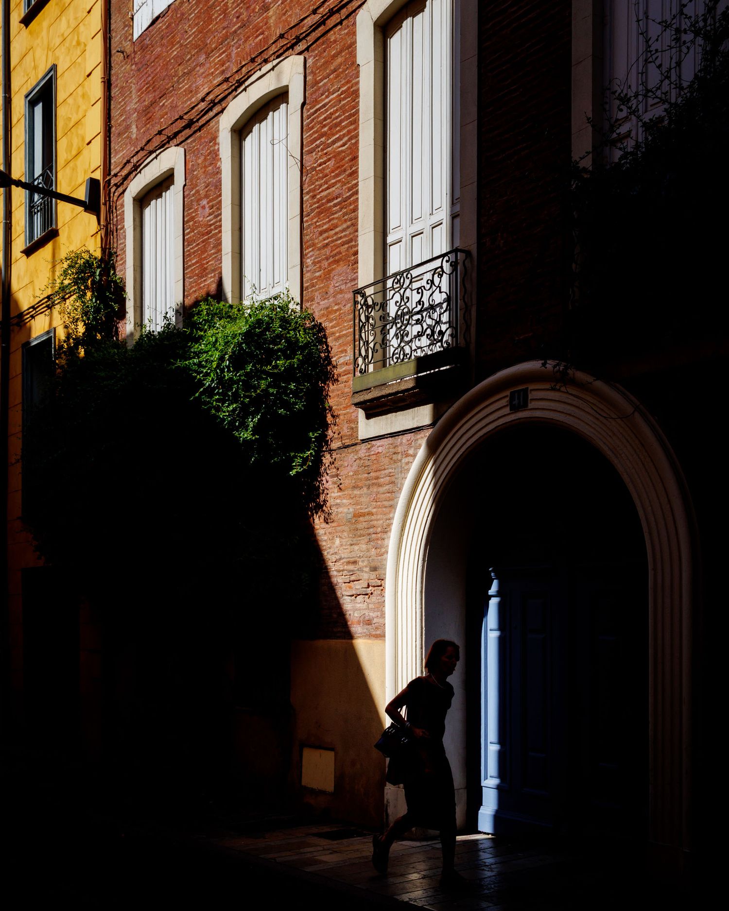 Silhouette of a person standing in a sunlit alley beside an ornate arched doorway in a historic brick building.