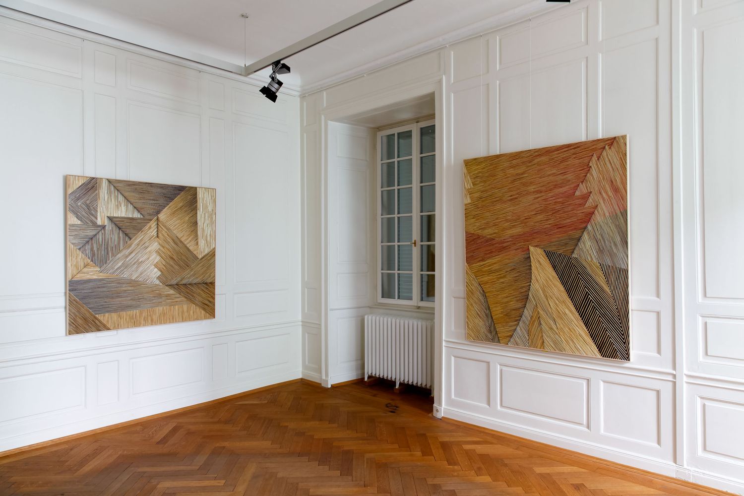 Gallery room with two large wooden geometric abstract artworks on white walls, herringbone parquet floor.