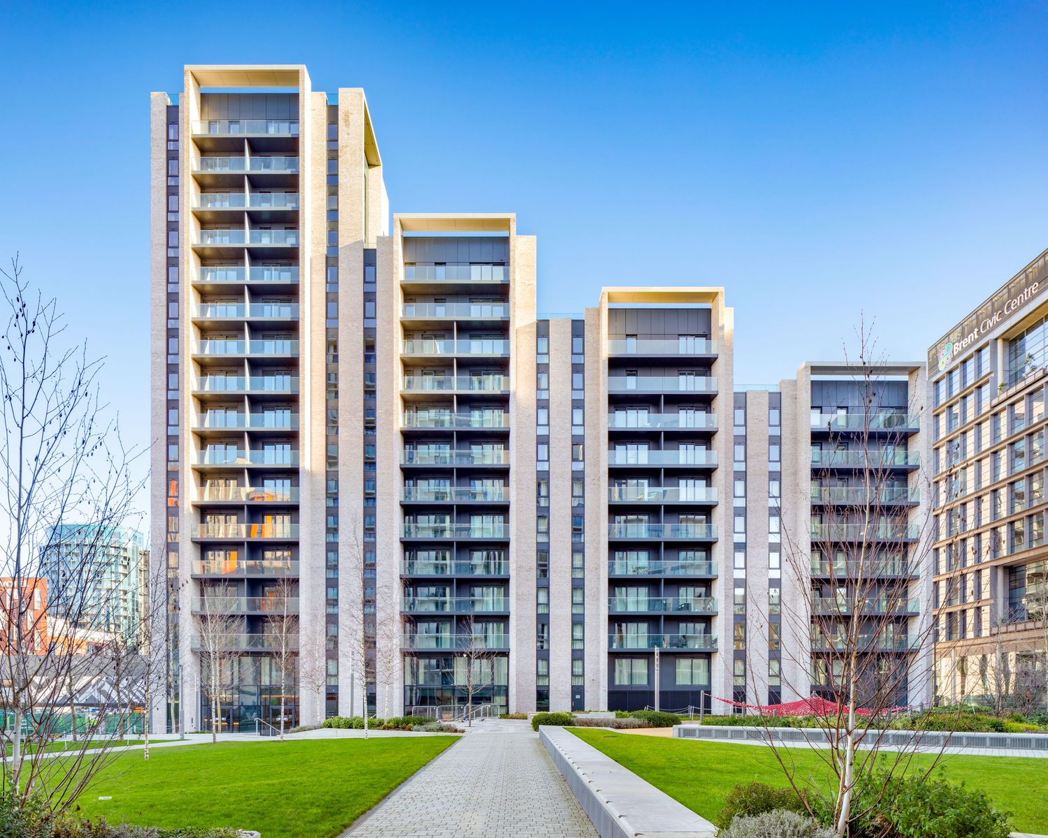 Modern high-rise residential apartment buildings with glass balconies surrounded by green lawns under a clear blue sky.