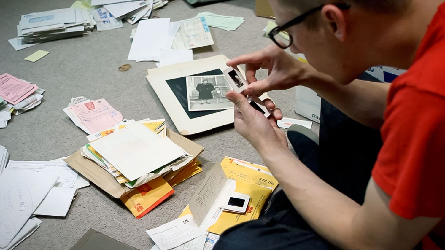 Man in glasses and red shirt examines an old photograph while sorting through scattered papers, folders, and documents on the floor.
