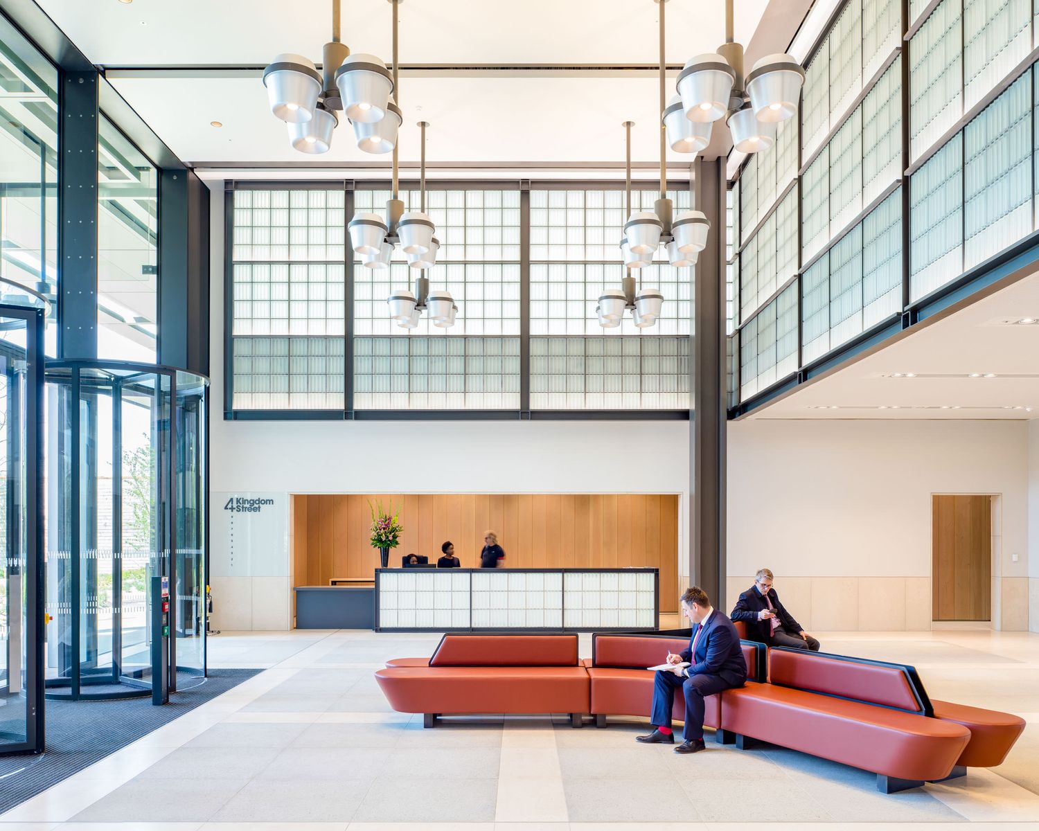 Modern corporate lobby with red modular seating, reception desk, globe pendant lights, and two professionals conversing.