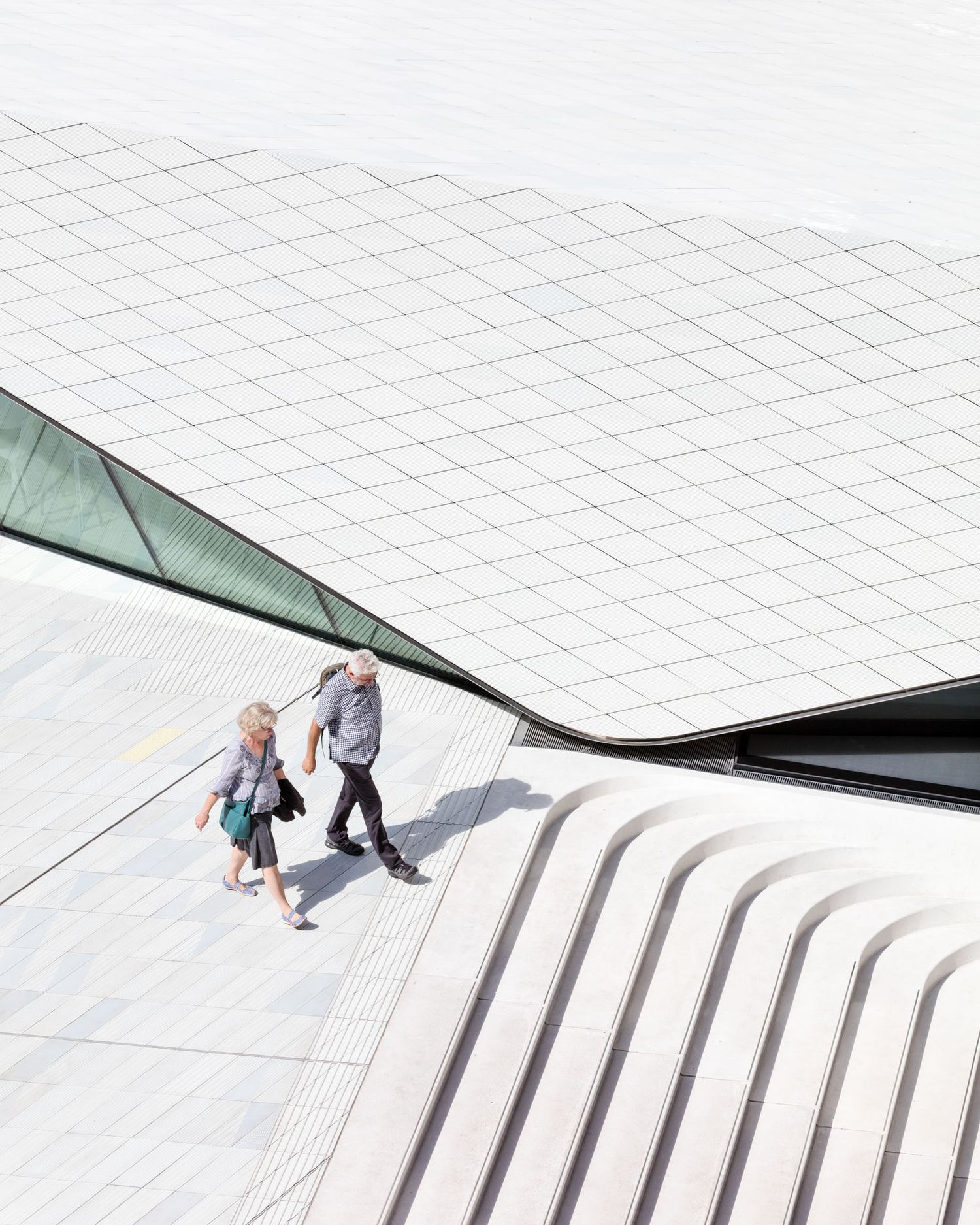Aerial view of two people walking beside a modern white tiled building with curved architectural lines.