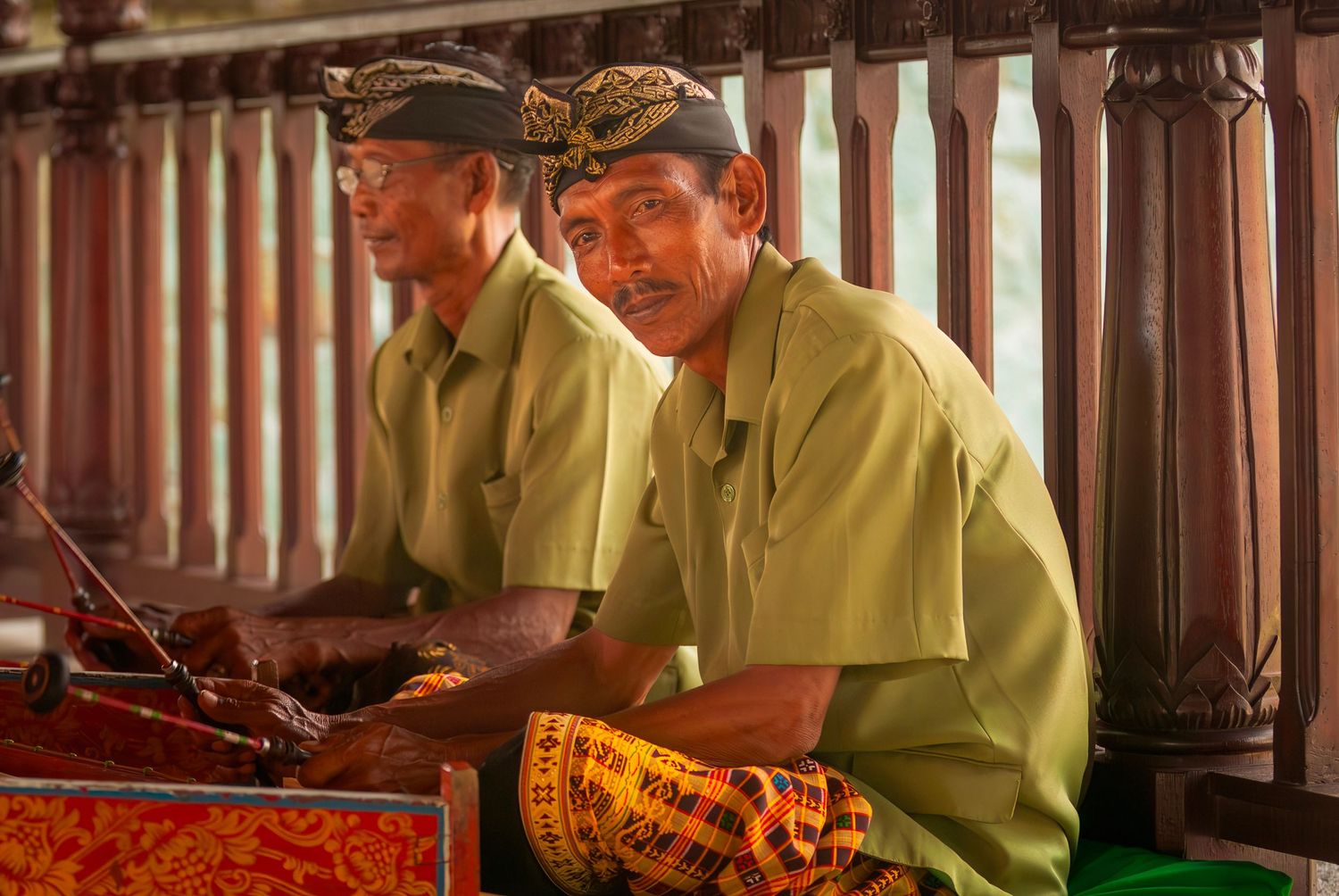 Two Balinese gamelan musicians in traditional attire playing instruments at a cultural performance in Bali.