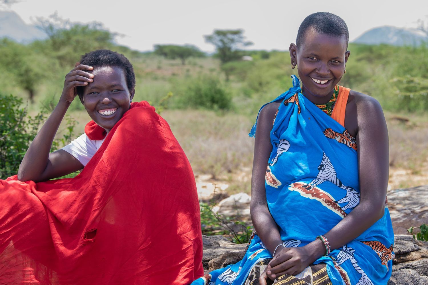 Two smiling Maasai women in colorful red and blue traditional clothing sitting outdoors in Africa.