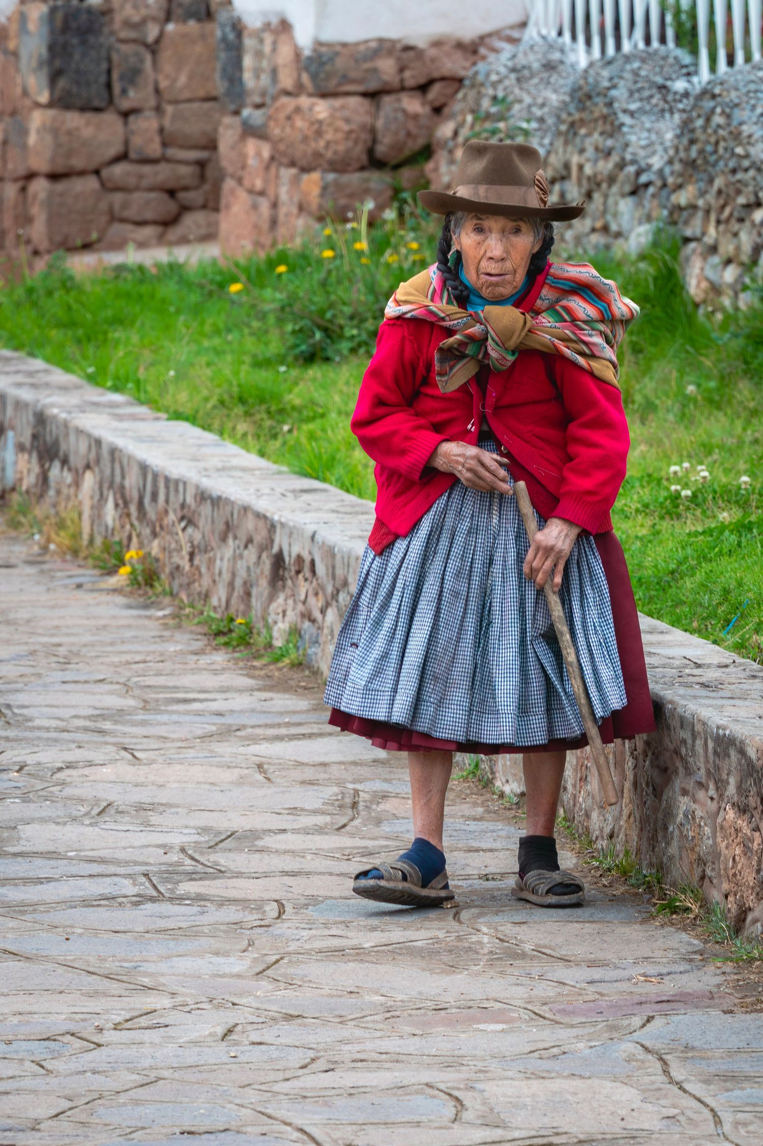 Elderly indigenous Peruvian woman in traditional red dress and hat walks with a wooden cane on a stone path.
