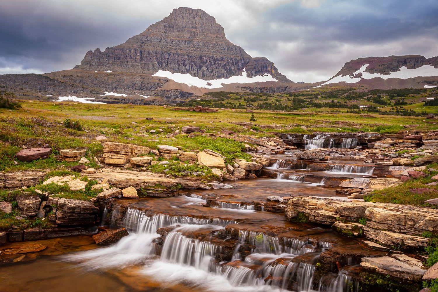 Scenic mountain waterfall cascades over rocky terrain in Glacier National Park under cloudy skies.