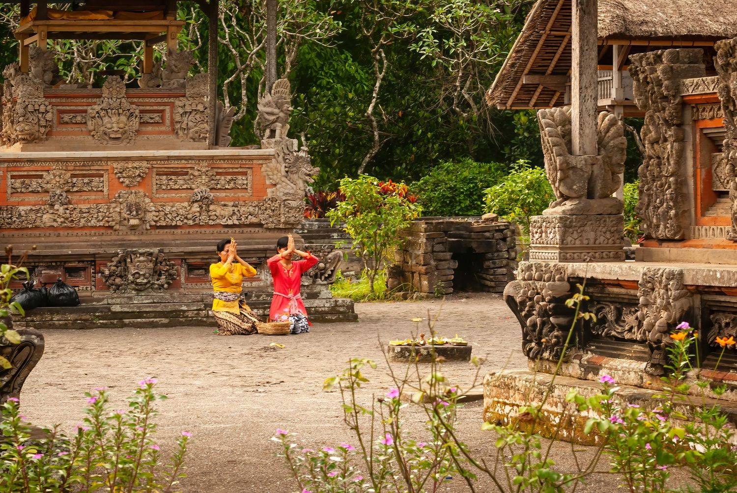 Two Balinese dancers in colorful yellow and red costumes performing in a traditional stone temple courtyard.